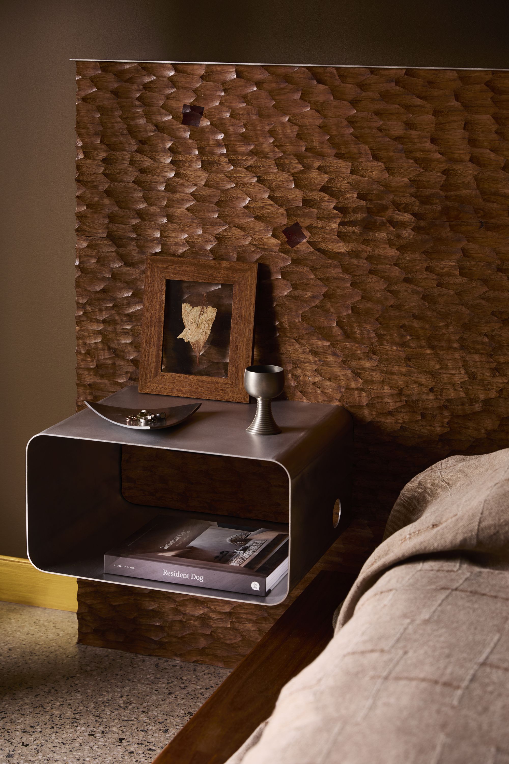 textured wood bedhead with floating stainless bedside table with book, goblet, picture frame, and polished concrete floor