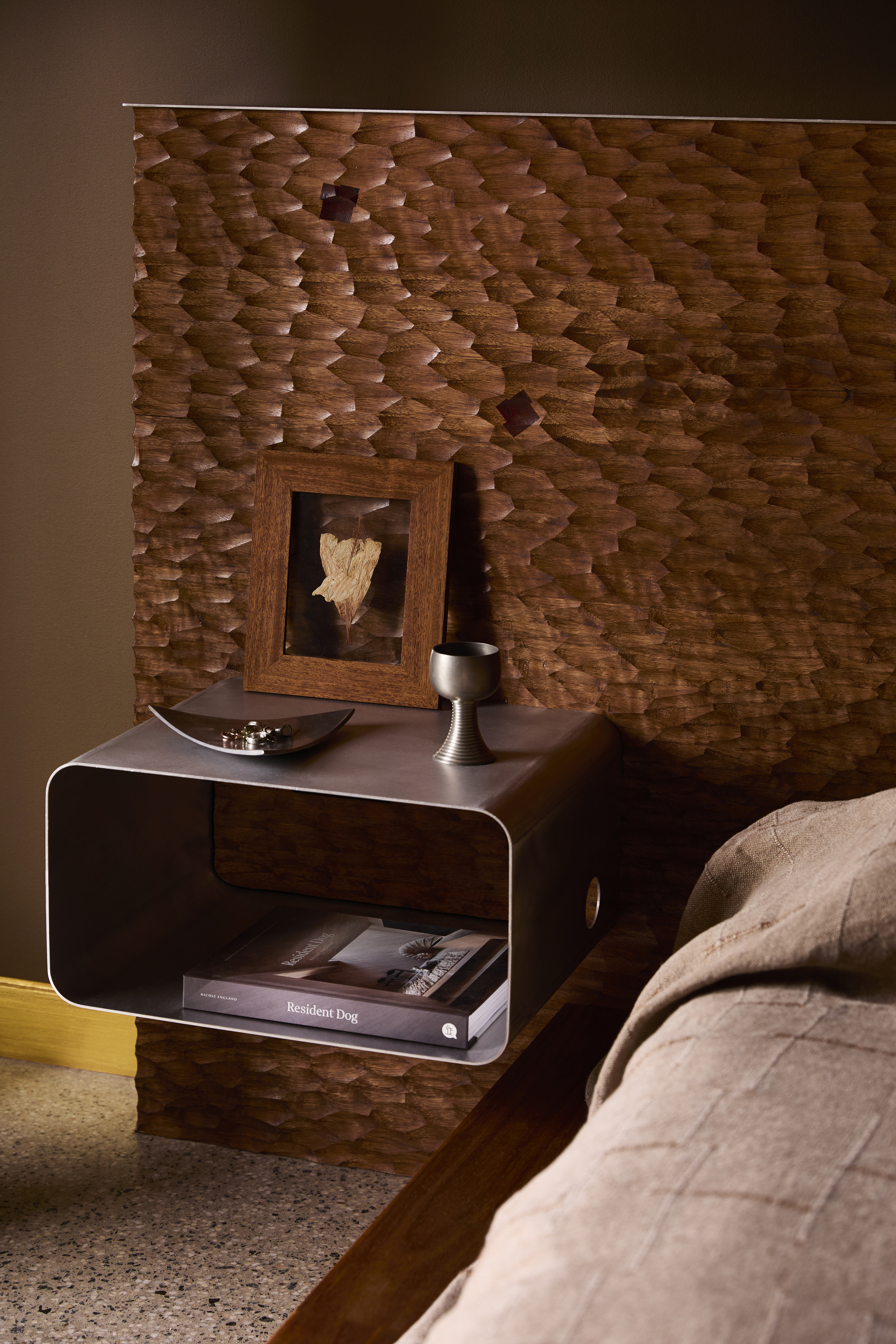 textured wood bedhead with floating stainless bedside table with book, goblet, picture frame, and polished concrete floor