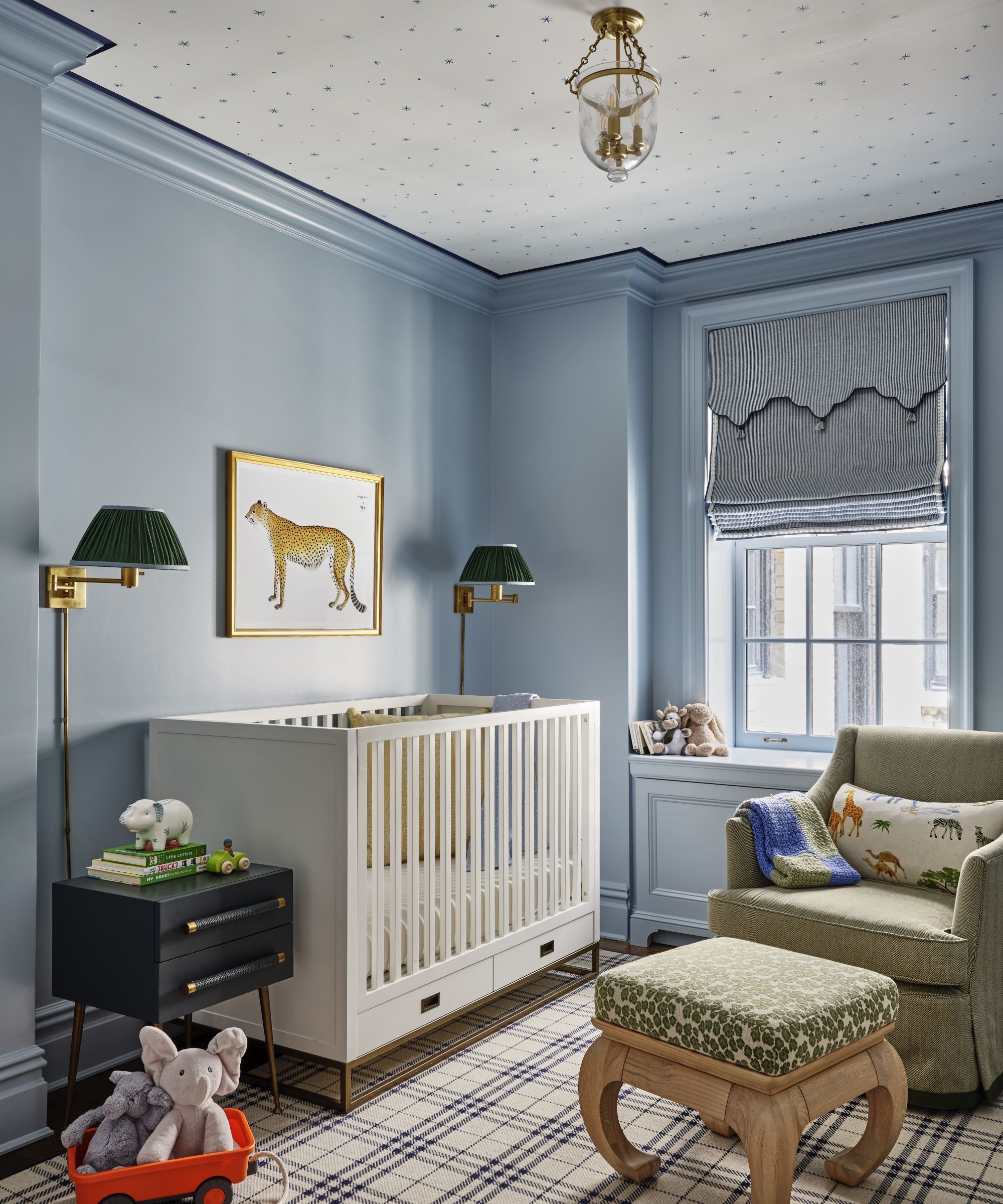 Nursery with pale blue walls, ceiling with hand-painted stars, white cot, two green sconces, an upholstered wooden stool, a green armchair and a plaid rug