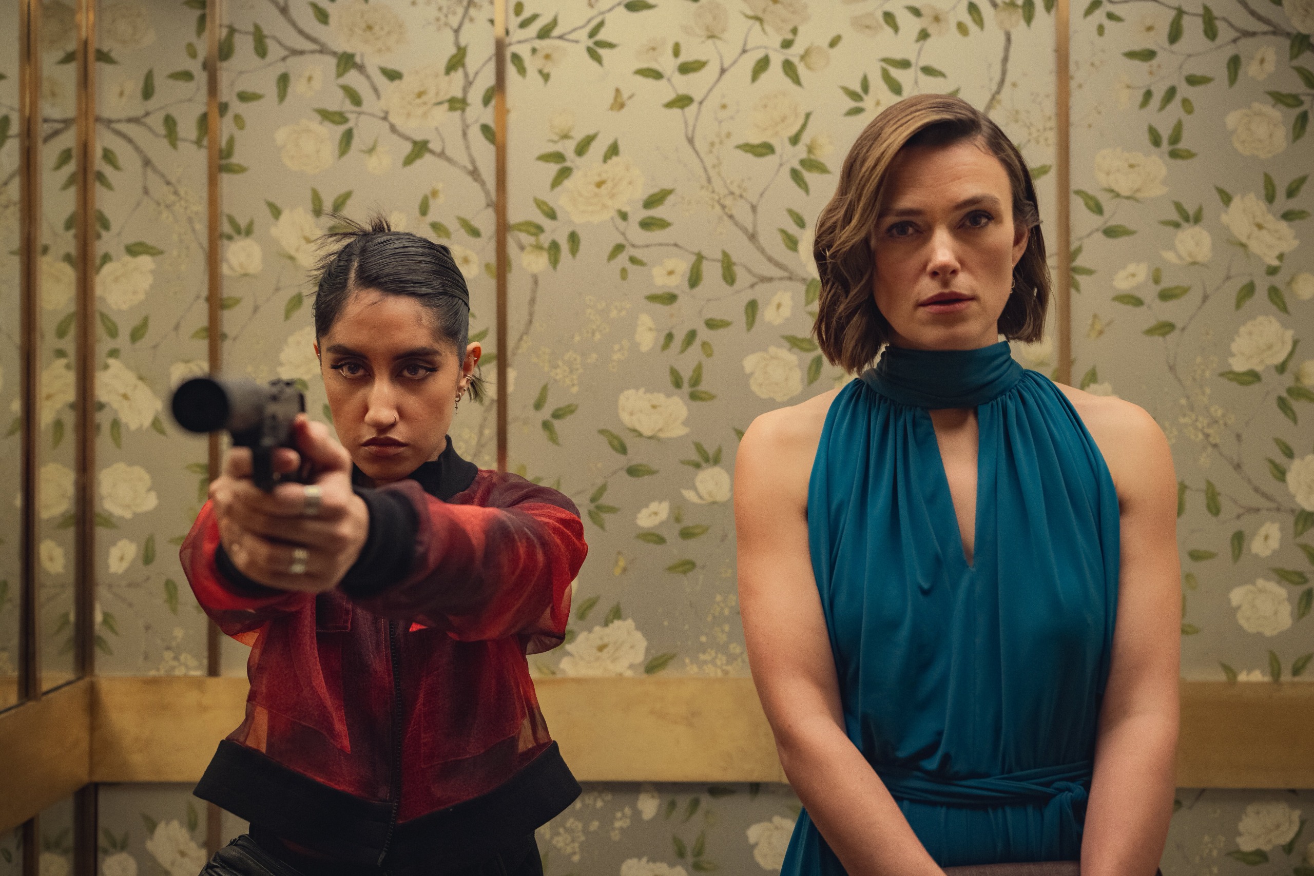 ambika mod holds a gun and keira knightley looks tense in an elevator decorated with flower wallpaper in season 2 of black doves
