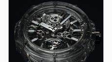 A clear plastic watch focussing on the see-through face. Under the face are the watch mechanisms. Two arms in silver point to 10 and 2. Hublot logo in the top part inbetween the watch arms and a mechanical feature. 