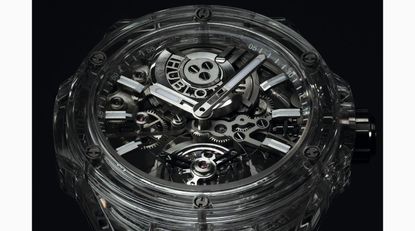 A clear plastic watch focussing on the see-through face. Under the face are the watch mechanisms. Two arms in silver point to 10 and 2. Hublot logo in the top part inbetween the watch arms and a mechanical feature. 