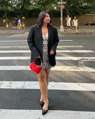 Female fashion influencer Marina Torres poses in a NYC crosswalk wearing an oversize black blazer, animal-print minidress, small red handbag, and black slingback heels.