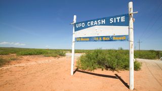 A sign reading "UFO crash site" stands on rusty red sand against a blue sky.