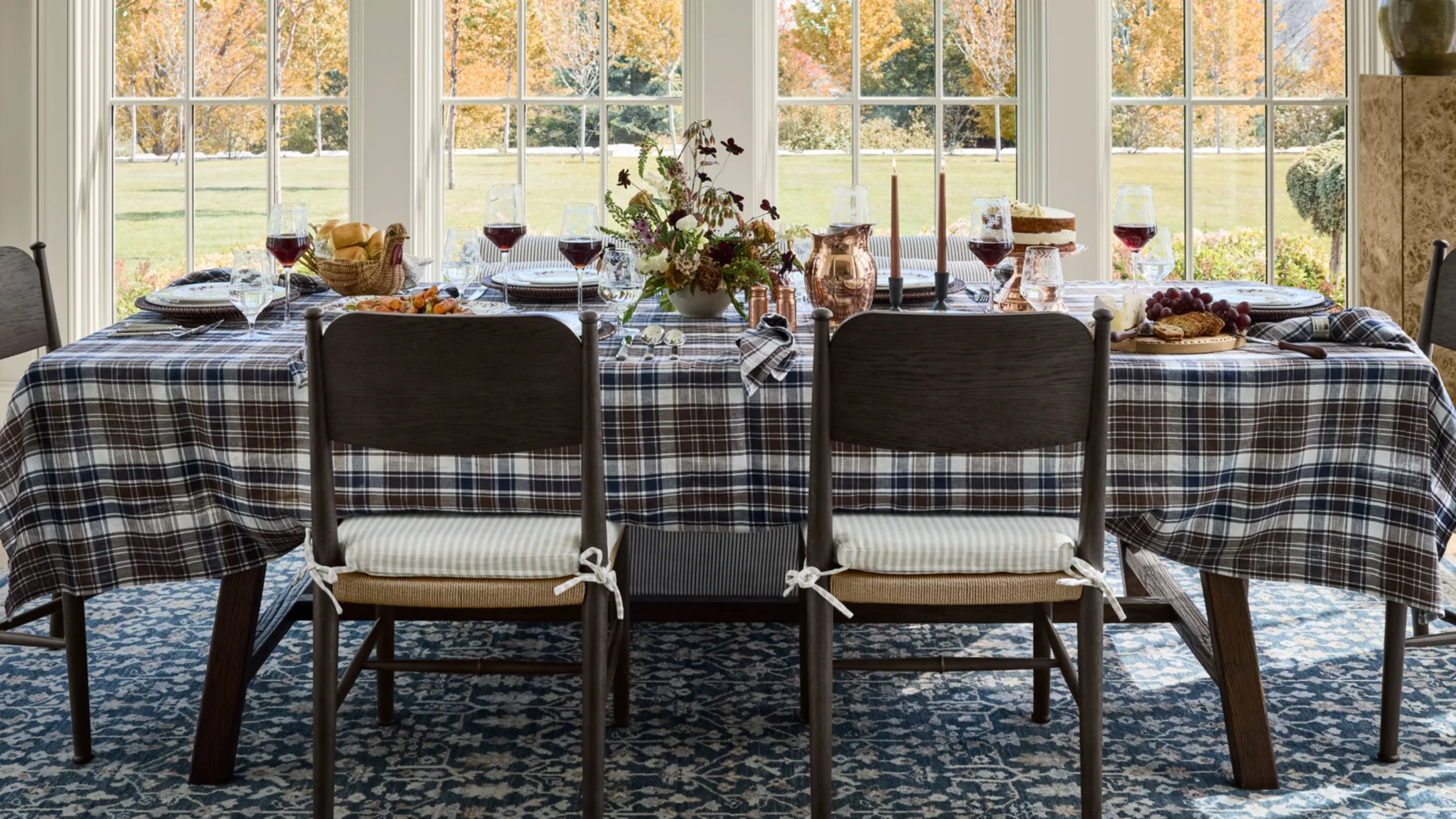 a fall tablescape with a plaid tablecloth, seasonal decor, and large windows in the background looking out to a large green field