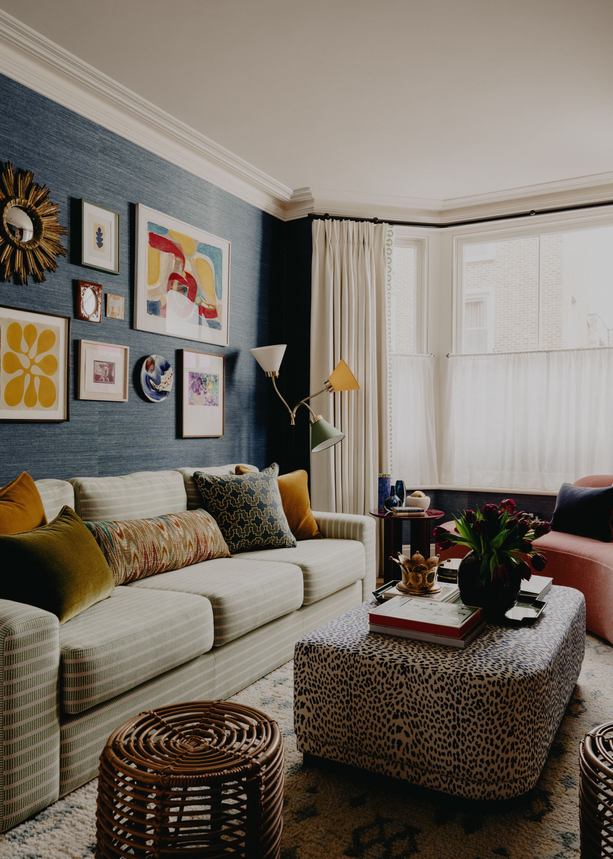 A cozy living room with dark blue walls, a gallery wall, a bay window with cafe curtains and cream curtains, a pale green striped sofa with colorful cushions, and a leopard print ottoman.