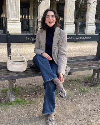 Parisian fashion influencer sits on a bench in Paris wearing a black turtleneck sweater, neutral blazer jacket, jeans, tan socks, and snakeskin ballet flats
