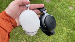A hand holding the white Sonos Ace and black Bose QuietComfort Ultra Headphones by their headbands with a grass lawn in the background.