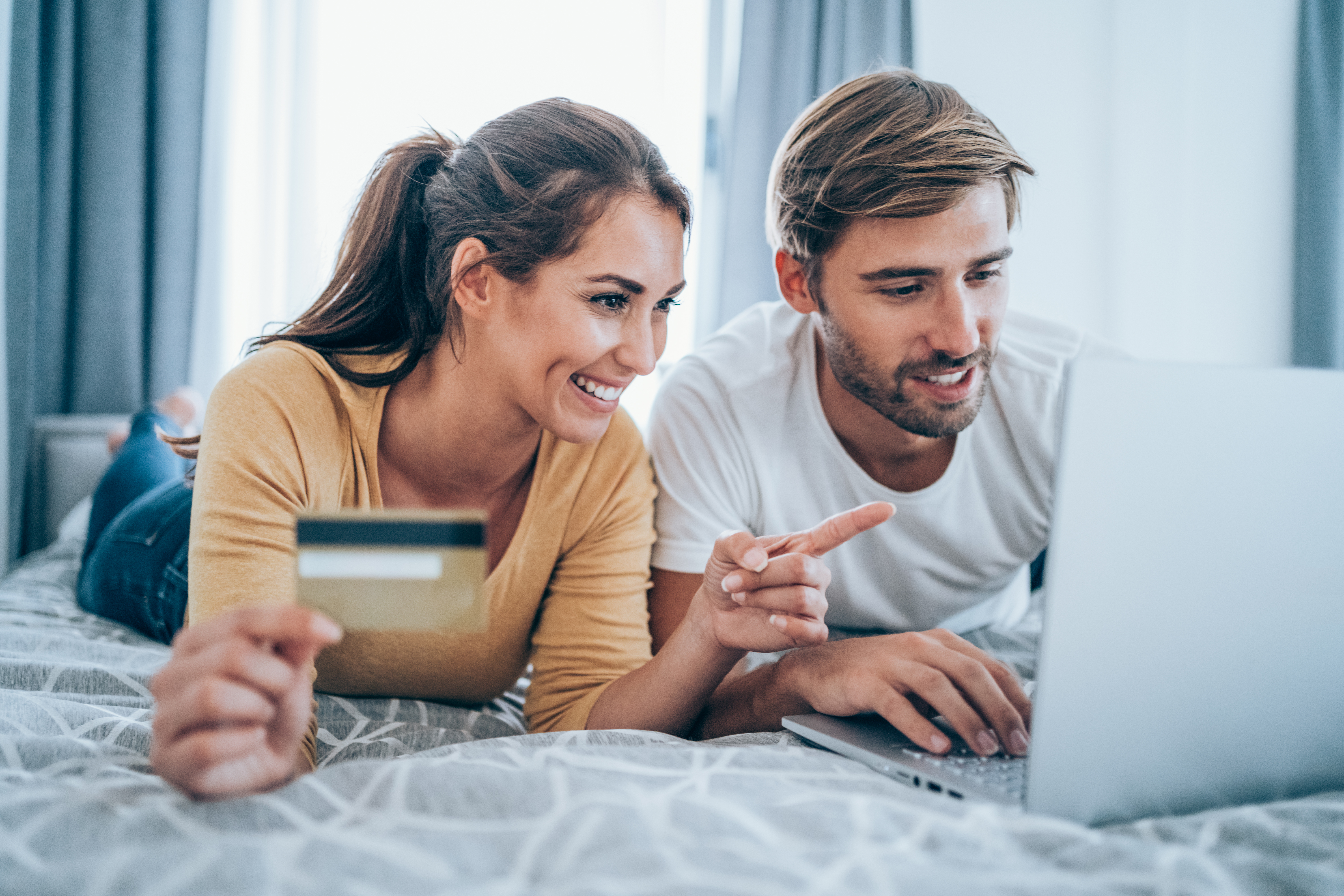 A couple using a laptop on a bed; one holds a credit card.