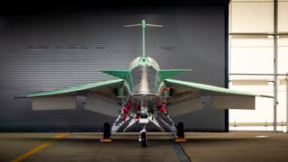 A head-on shot of NASA's X-59 supersonic jet, whose wingspan stretches across the middle of the photo. Green and black panels of the jet's body reveal sleek, sharp angles. The jet sits on three wheel connected by landing gear struts. The jet sits inside a hanger with white walls, and a grey corrugated door closed behind the jet.