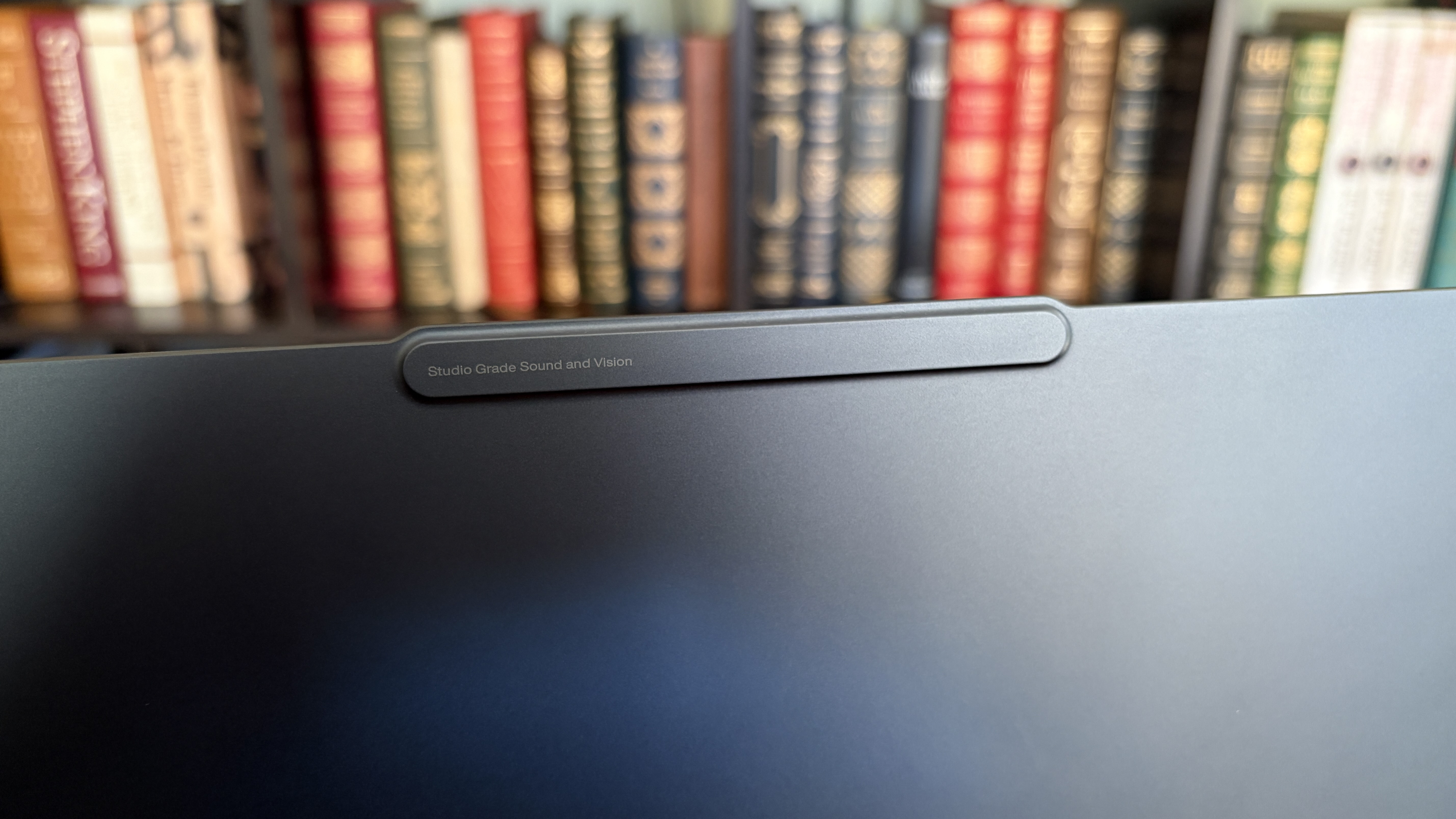 Close-up of a computer monitor's top edge with &amp;quot;Studio Grade Sound and Vision&amp;quot; text, against a blurred background of colorful books on a shelf.