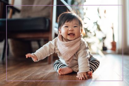 Asian baby girl wearing baby bib with a cheeky smile, sitting on the floor in the living room. Baby development milestones.