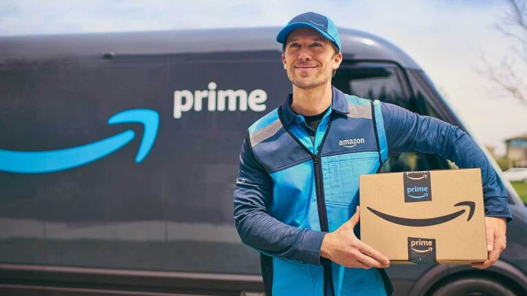 An Amazon Prime delivery driver in branded uniform holding packages while standing in front of a Prime-branded delivery van.