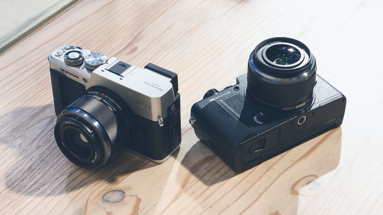 Two OM System PEN cameras on a wooden table, one in silver one black