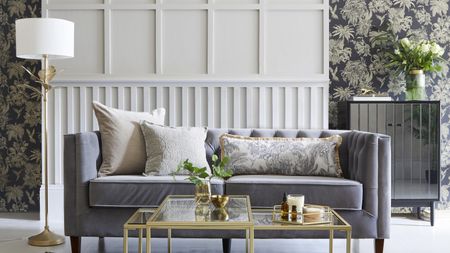 A white and grey living room with white wall paneling, charcoal grey and gold palm-print wallpaper, grey velvet sofa and palm print rug 