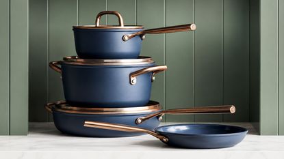 A stack of blue pans with brass effect handles and lid rims, on a marble kitchen counter with green background.