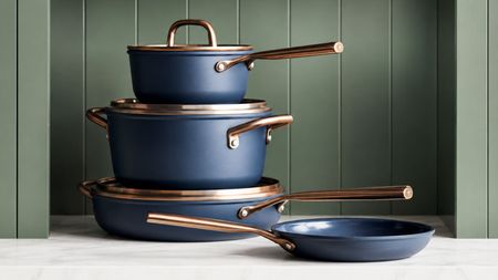 A stack of blue pans with brass effect handles and lid rims, on a marble kitchen counter with green background. 