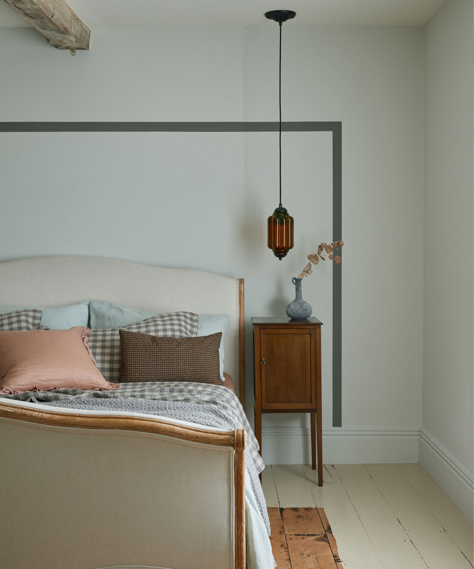 Bedroom with a rustic touch, featuring a light wooden bed frame and a mix of soft, checkered and plain cushions. A vintage amber pendant light hangs beside a simple wooden bedside cabinet, creating a balanced blend of traditional and modern design