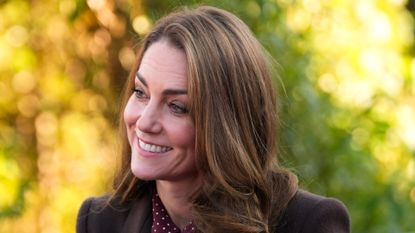 Kate Middleton wearing a brown coat looking to the left and smiling in front of a background of gold and green fall leaves