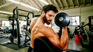 Shirtless man performs dumbbell biceps curl using angled preacher bench to rest his upper arm on