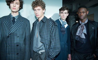 Four male models wearing wearing suits and trousers in shades of blues and black, raglan-sleeved trenches, complete with exaggerated epaulets, which were teamed back with Shetland wool crew neck sweaters and loosely cut trousers, finished with pussy-bow tied fringed scarves.