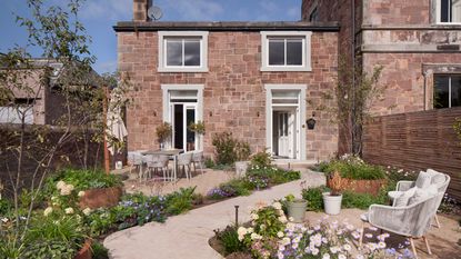 a house with garden in the foreground with a winding pathway, two seating areas and lots of flowers in purple and white