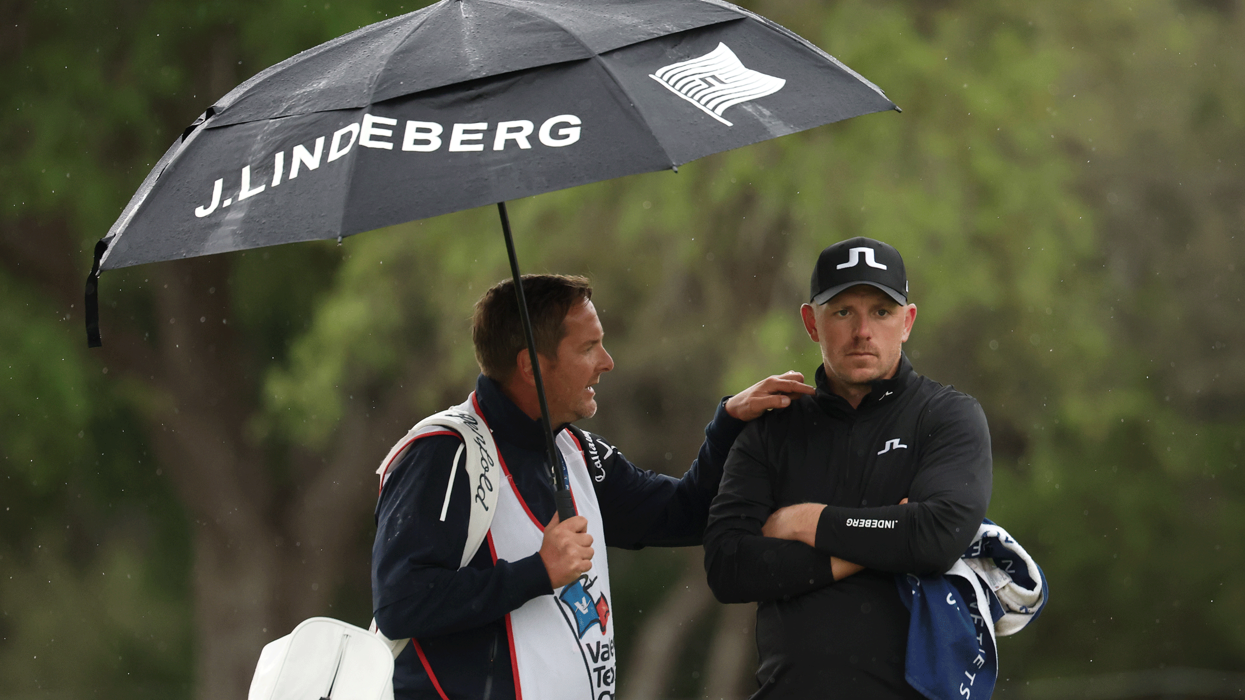 Matt Wallace looks on during the final round of the Valero Texas Open while his caddie talks to him while putting his hand on Wallace's shoulder and holding an umbrella over their heads