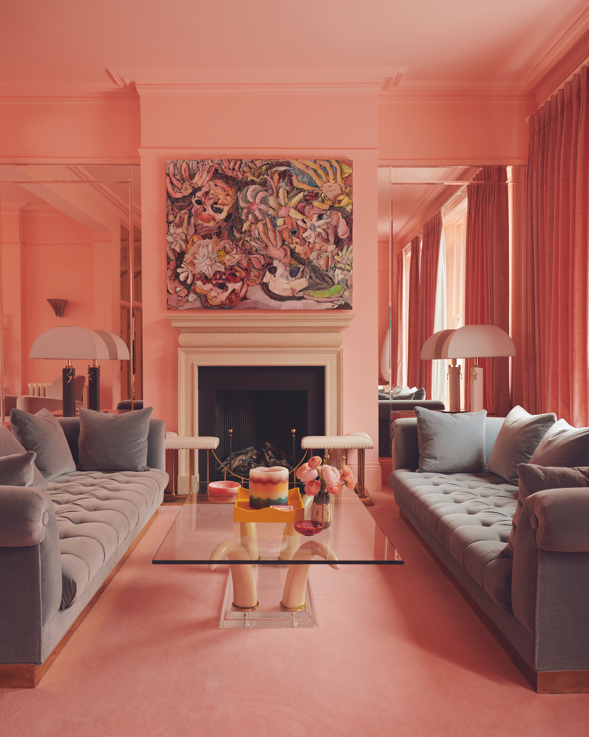A coral pink, color drenched living room, with grey sofas facing each other, a glass coffee table in between and an artwork hanging above the fireplace, with mirrored alcoves and lamps in each.