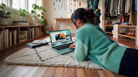 A woman lying on a rug in a modern room using a teal Dell 14S laptop running Windows 11.