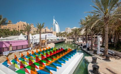 Outside daytime image, clear canal water, colourful transparent triangles on a floating white platform art piece, palm trees, canal bank, bars and tall buildings in the backdrop, clear blue sky