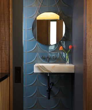 A small bathroom with dark blue scalloped walls, a circular hanging mirror, a marble sink, and a vase of orange flowers