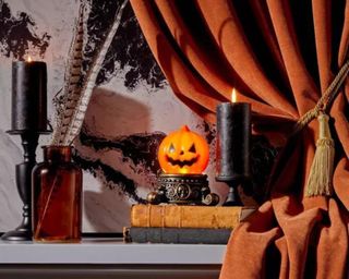 A shelf with orange and black books, candles, an orange pumpkin, and a dark brown curtain on top of it