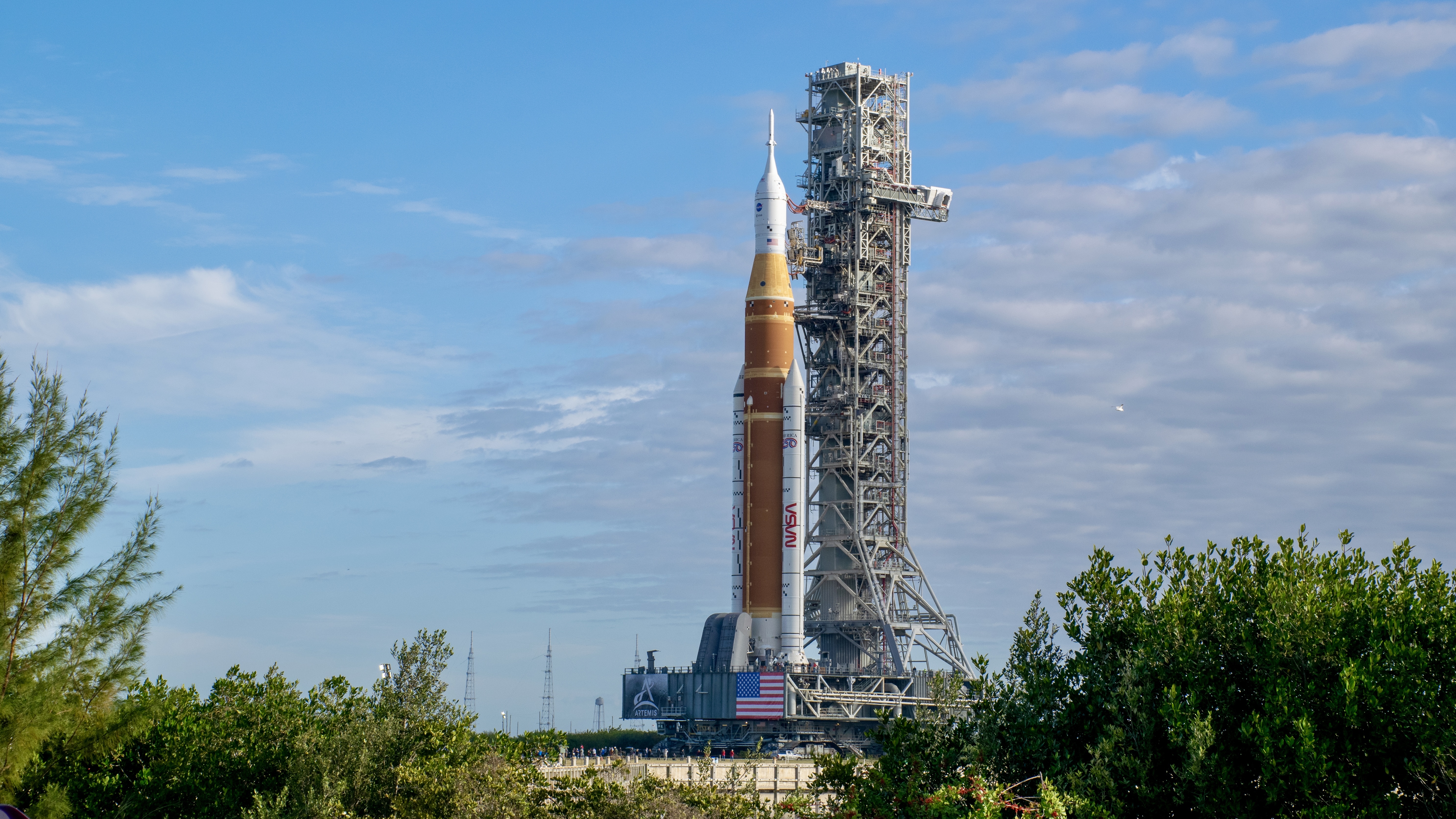 A big orange rocket stands in the distance next to a launch tower. On the bottom left and right are green bushes in the foreground.