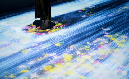 Art installation of TeamLab in the Pace Gallery. Neon, glowing colors cover the floor. We see a bottom portion of a woman standing in the middle of it, with a black space around her feet.