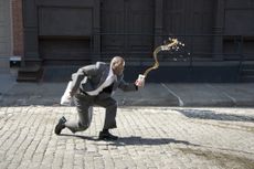 A man in a business suit trips and spills coffee outside on a cobblestone street.