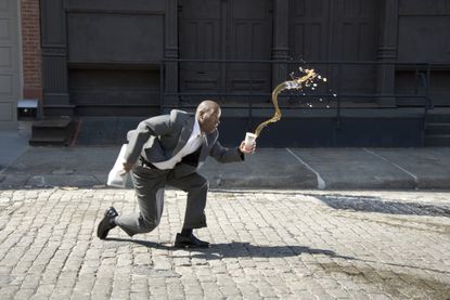 A man in a business suit trips and spills coffee outside on a cobblestone street.