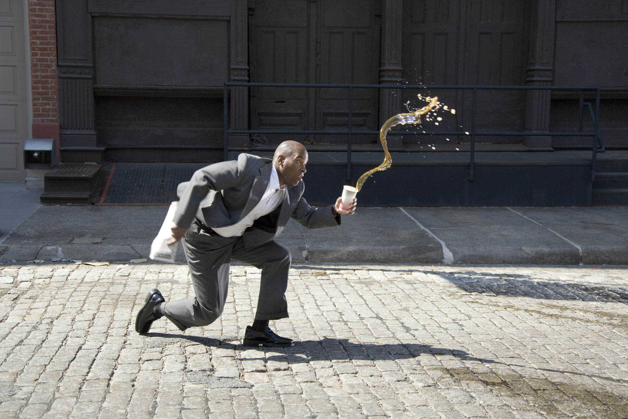 A man in a business suit trips and spills coffee outside on a cobblestone street.