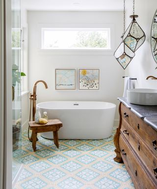 narrow bathroom ideas, patterned tiles on floor, bath, vintage stool and sideboard, brass standpipe, glass pendants