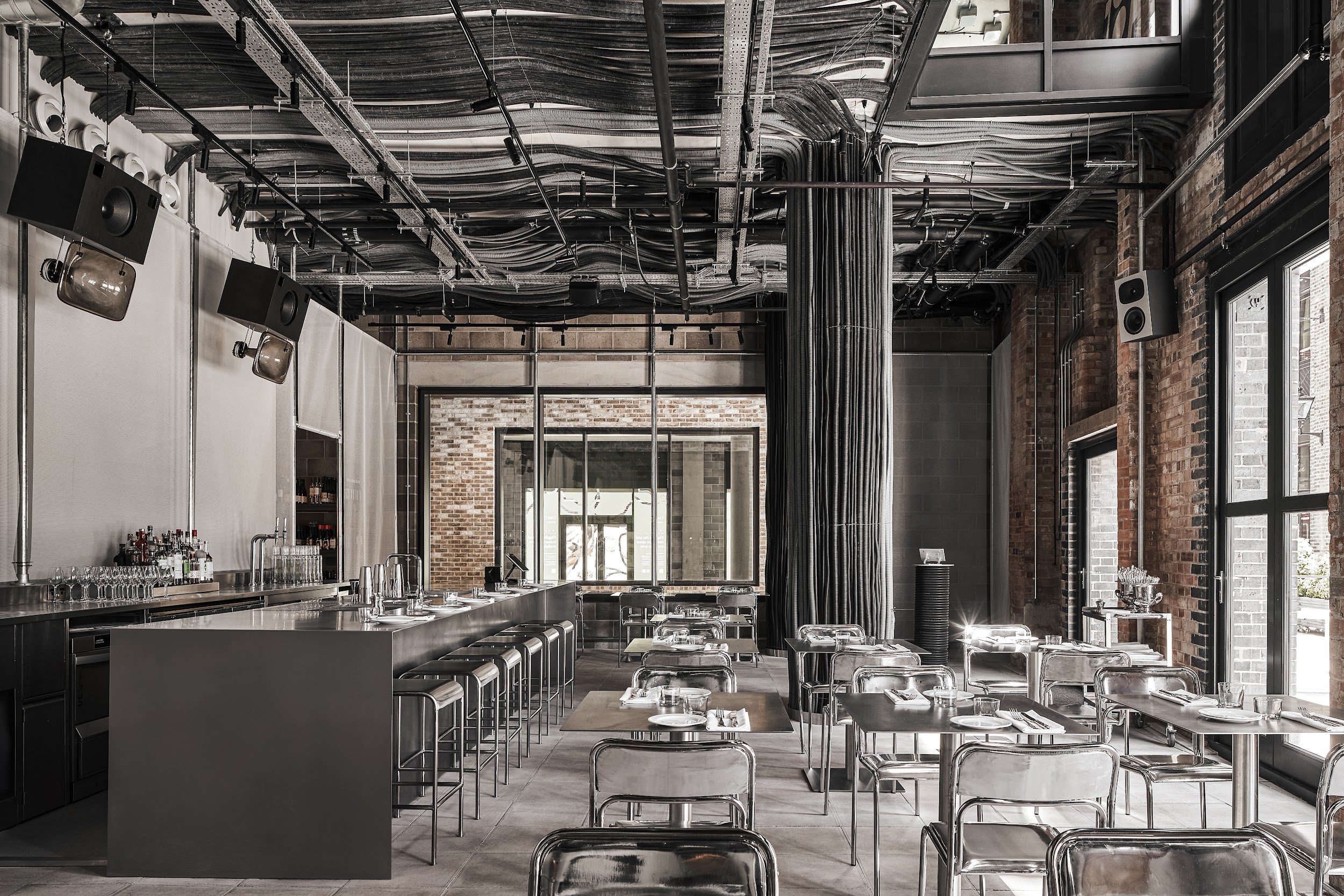 An industrial-chic restaurant features an exposed ceiling draping in wires, ventilation pipes, and cables, white cement or brick walls, and monochromatic chrome table, chair, and bar designs.