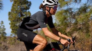 A cyclist wearing apparel from Le Col's ARC Gravel Clothing range cycles past some trees