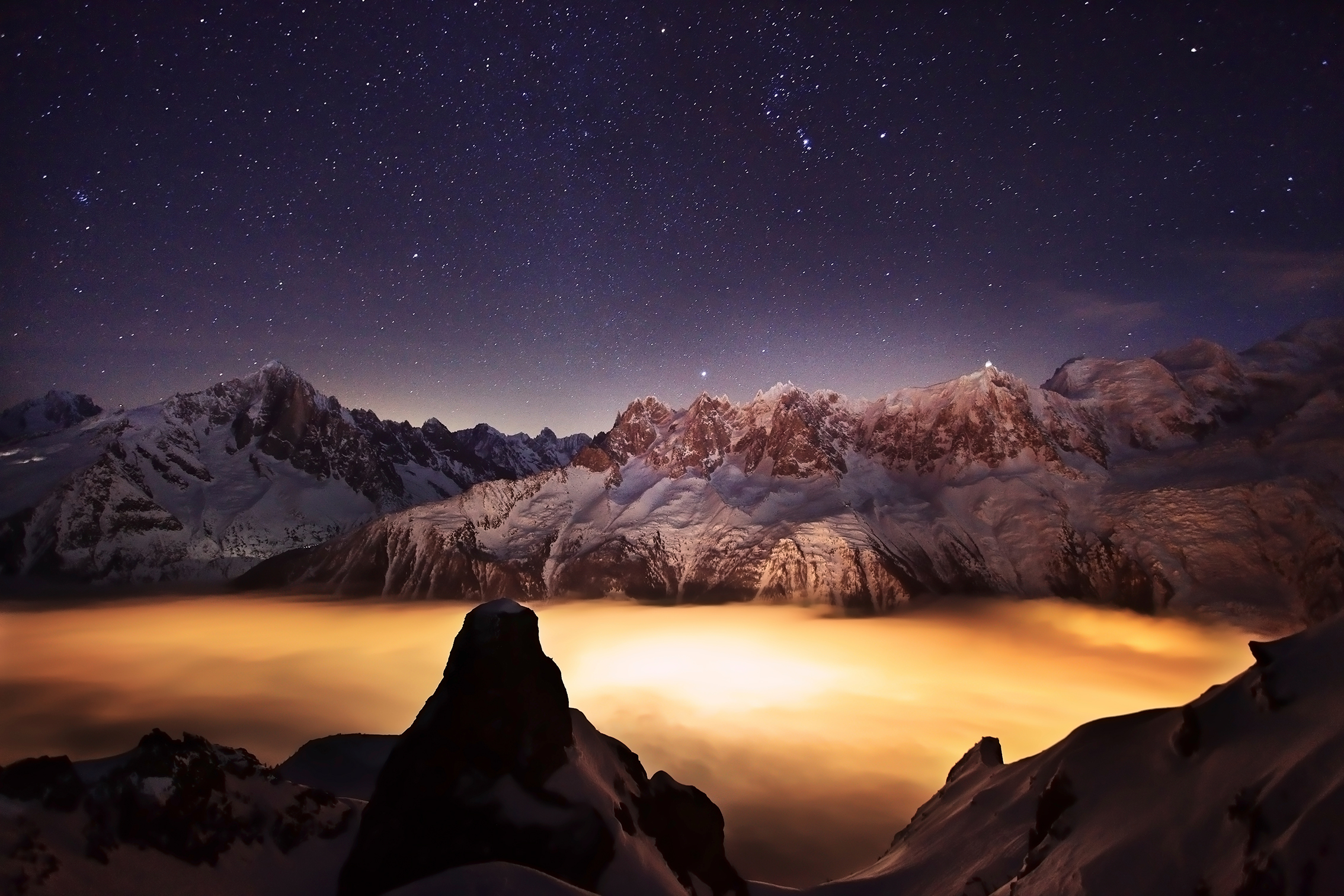 A stunning night landscape of snow-covered mountains under a starry sky, with glowing clouds illuminated by distant lights