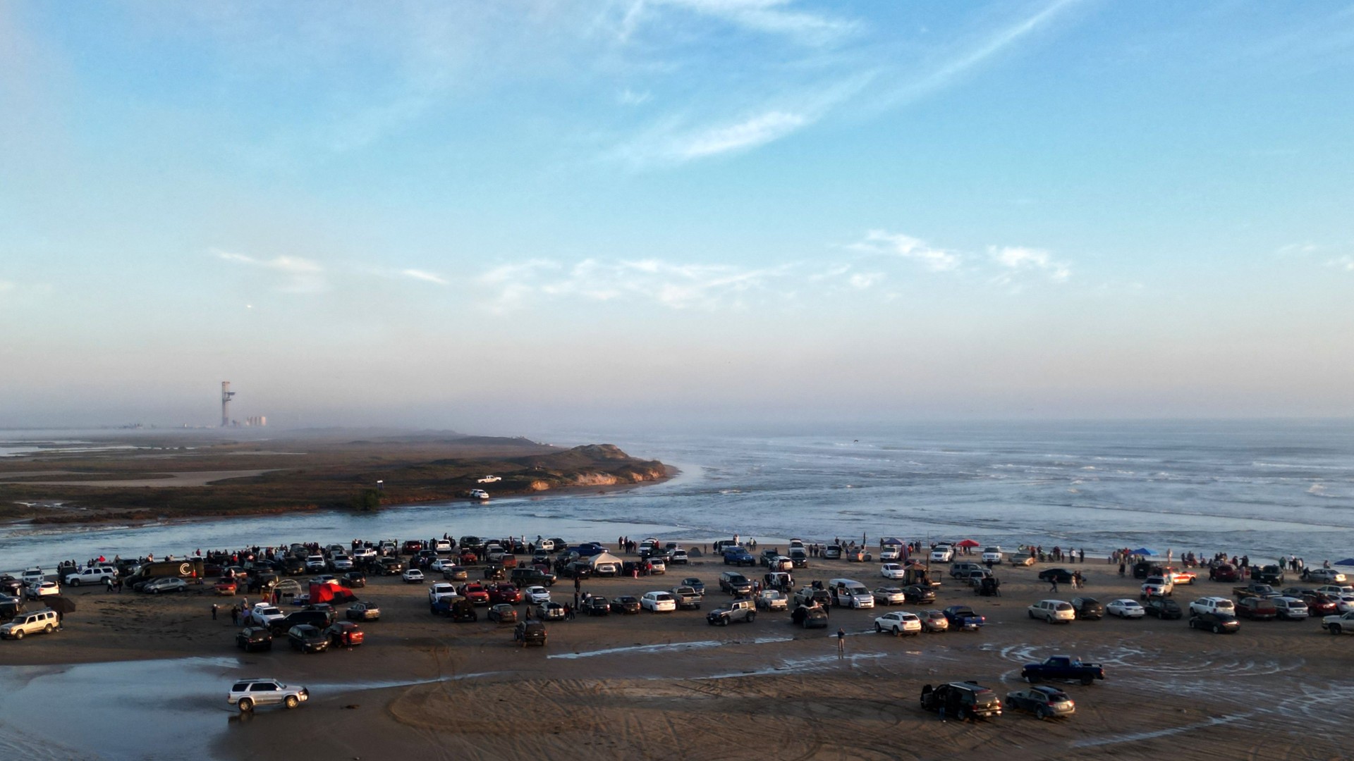 cars and onlookers line a beach in the foreground, with a large metal tower visible in the background
