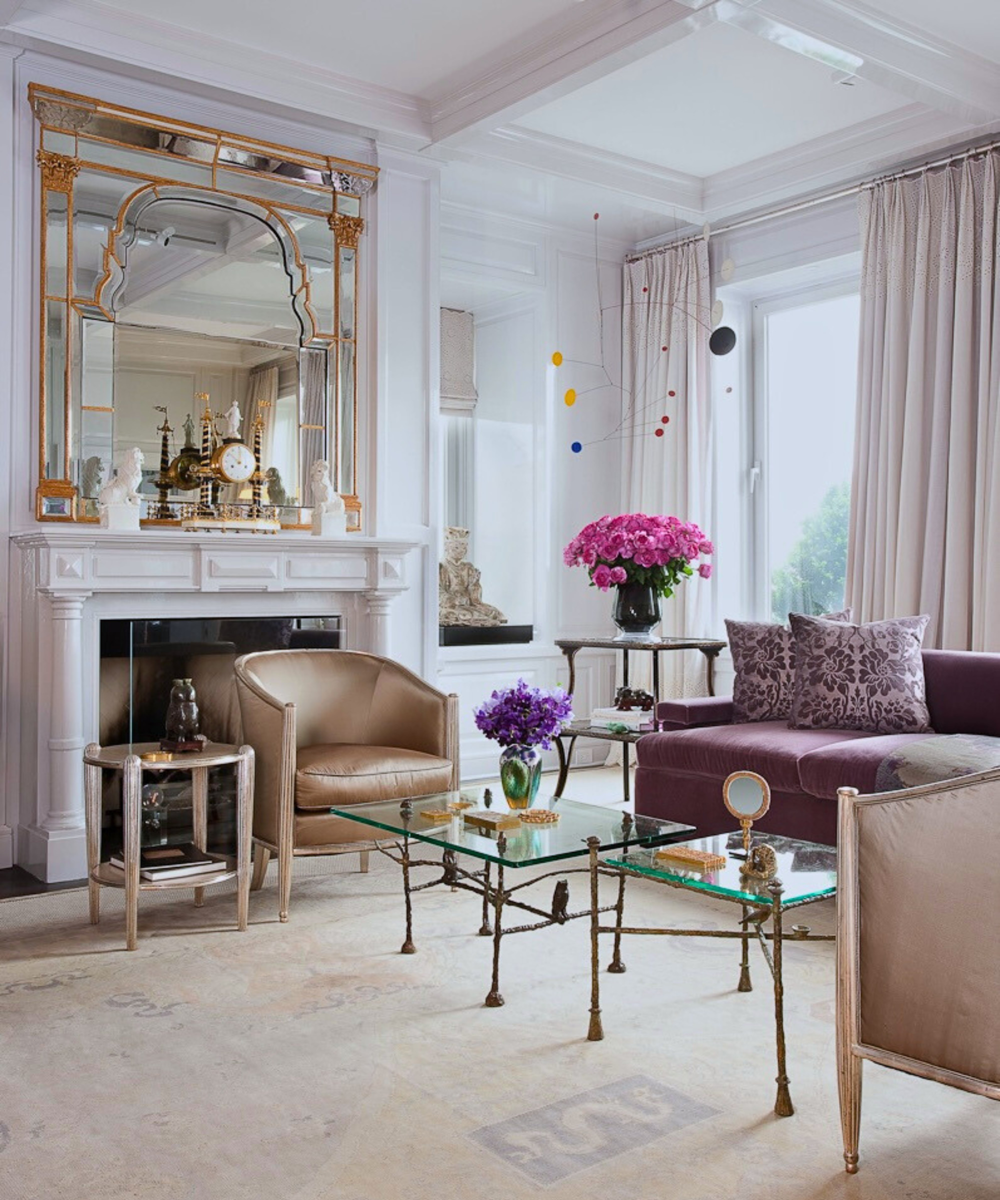 A bright living room with high ceilings, a white fireplace, glass coffee tables and tan armchairs