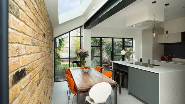 a kitchen extension by PlusRooms, with a large white island and white counters, hanging lights, a wooden dining table and a brick wall, to illustrate the extension cost calculator