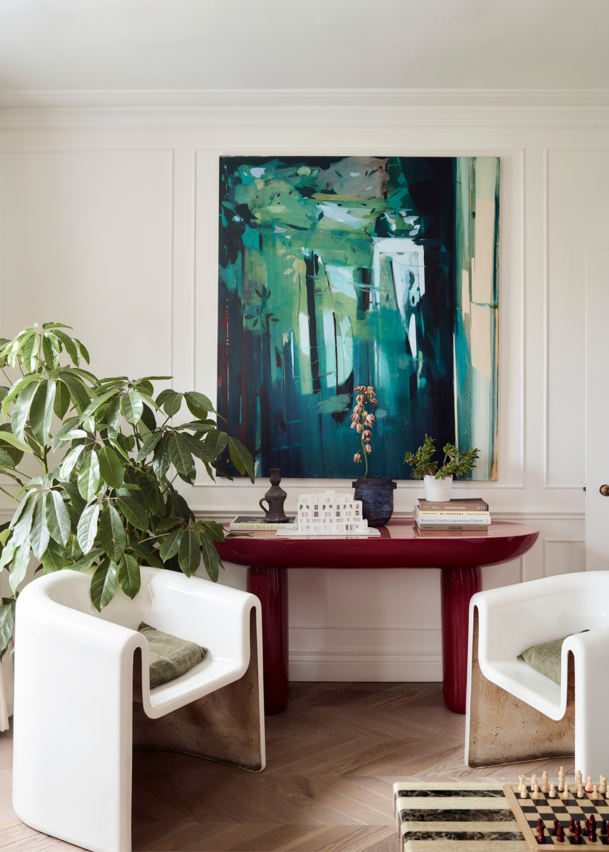 A living room corner with a colorful blue painting, a potted plant, a pair of white armchairs, a red side table with a stack of books, potted plants, and a peak of a stone coffee table with a chess board