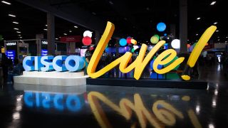 A sign with the word Cisco written in blue next to the word live! written in yellow, both of which are sat on a dark conference room floor
