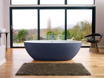 Bathroom image, wooden floor, blue and white free standing bathtub, brown rug, black framed window with view of the surrounding garden. black chair, black side table