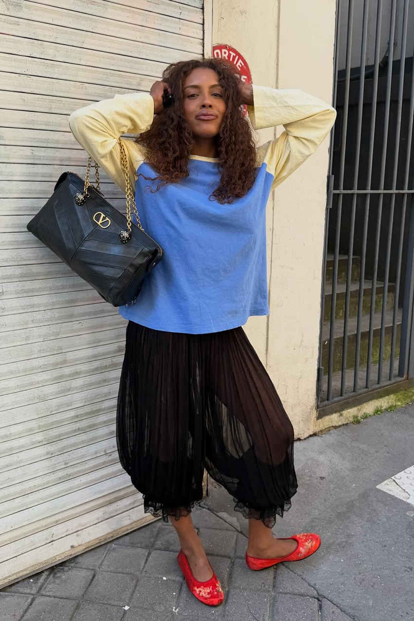 a woman wearing a yellow and blue shirt, sheer balloon pants, and red flats