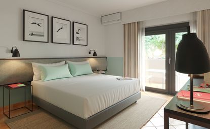 Interior view of a room at The Magnolia Hotel featuring light coloured walls, a grey bed base with white and pastel green pillows and white linen, black wall lamps and framed wall art. There are also black bedside tables, a light coloured rug underneath the bed, a table opposite, a glass window and door with a grey frame and light coloured curtains