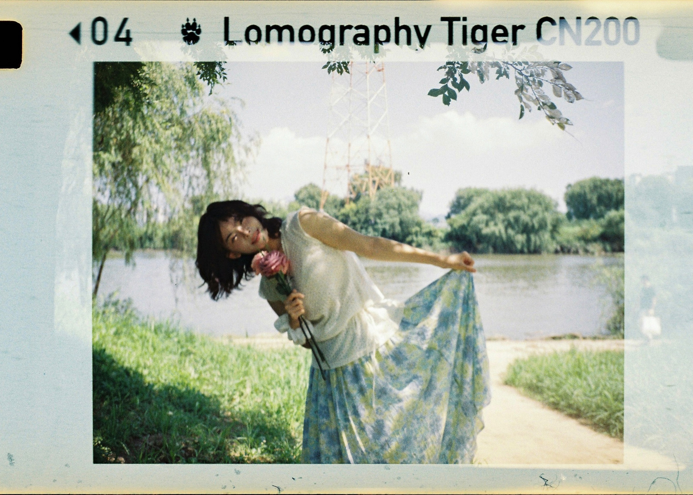 Photo of a woman in a sleeveless top and floral skirt holding flowers, posing by a lake with trees and a radio tower in the background